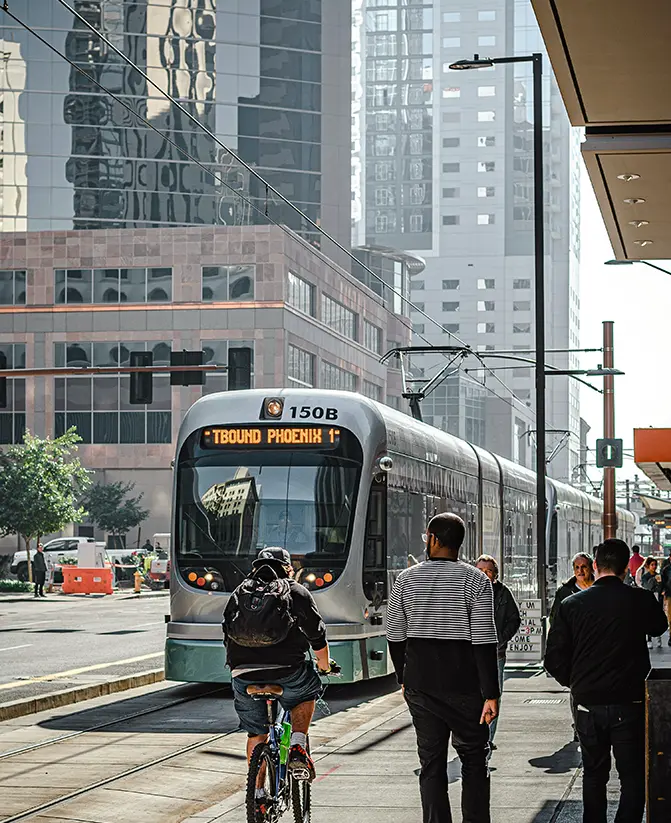 A modern light rail train with a “TBOUND PHOENIX” sign approaches a station on a bustling city street, surrounded by tall reflective buildings.